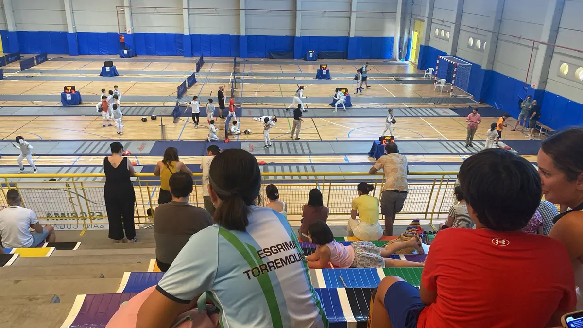 Pabellón con varias pistas de esgrima, niños compitiendo y familias en la grada, una con la camiseta del club