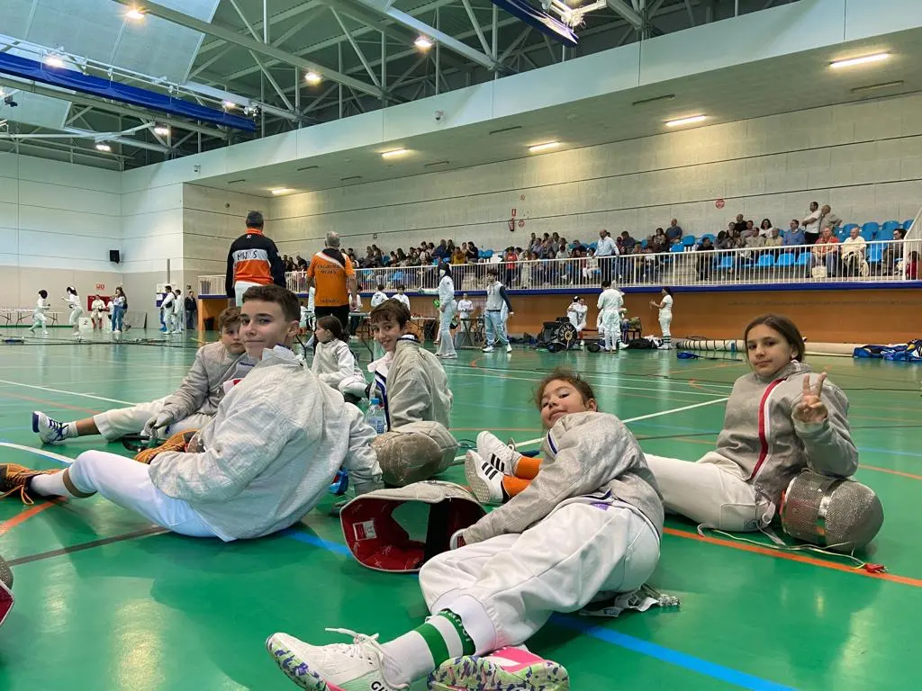 Niños y niñas esgrimistas sentados en la pista con sus caretas durante una competición