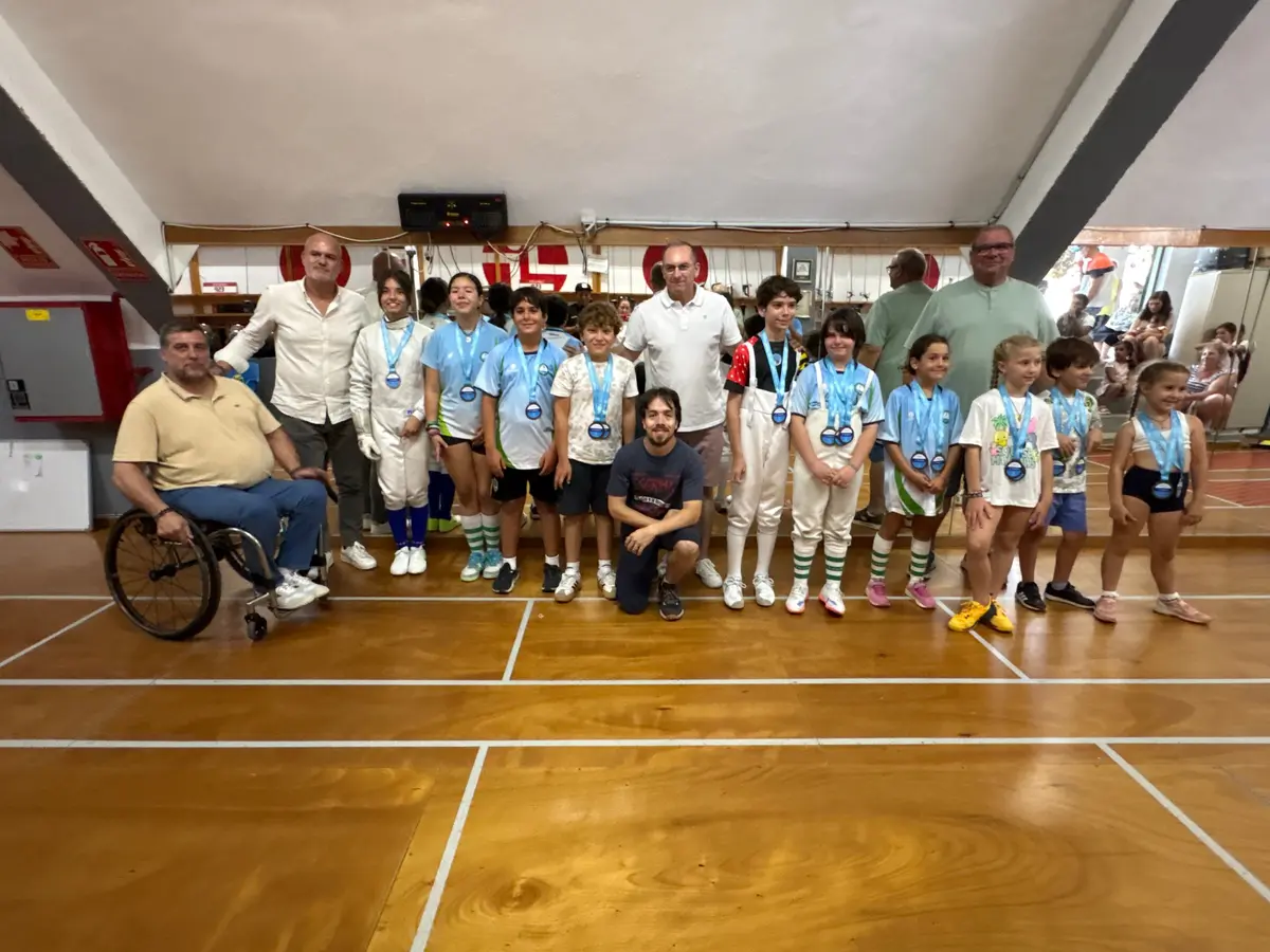 Grupo de niños y niñas con medallas azules posando junto a adultos, uno de ellos en silla de ruedas, en la sala del club
