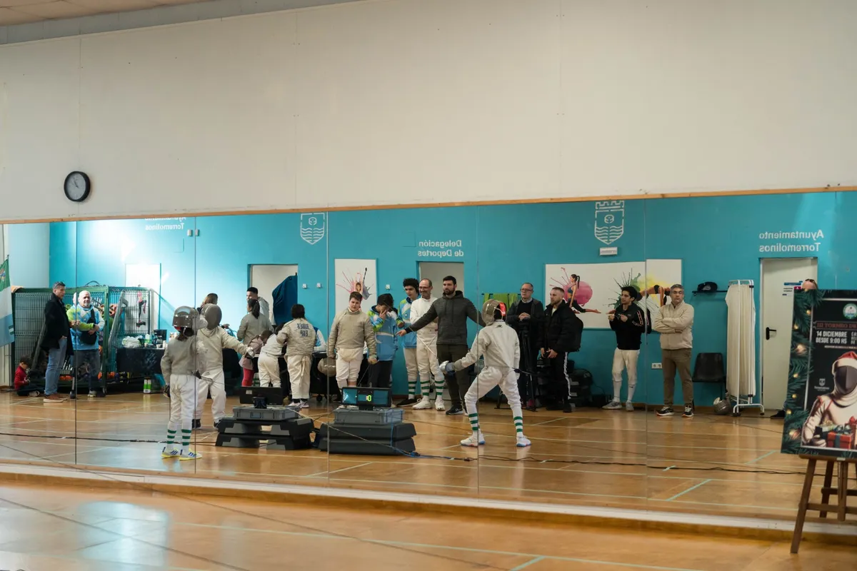 Asaltos de sable reflejados en el espejo de una sala municipal de Torremolinos, con familias y entrenadores mirando y el cartel del Torneo de Navidad