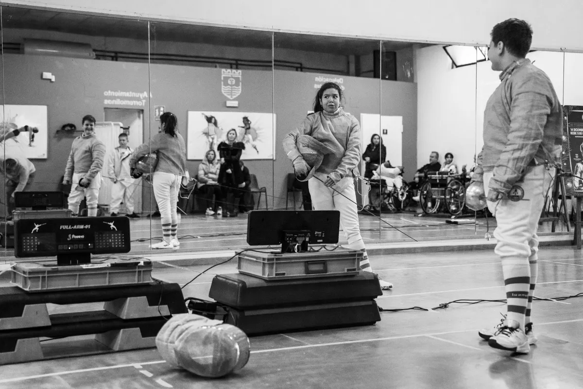 Fotografía en blanco y negro de dos jóvenes tiradores de sable entre asaltos, junto a los marcadores y el espejo de la sala